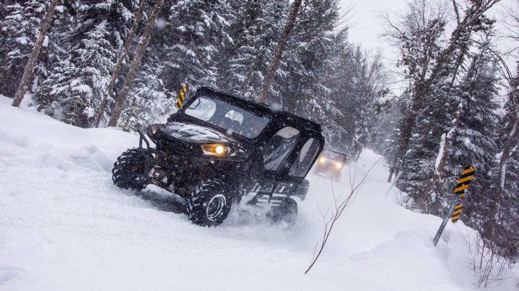 Two off-road utility vehicles drive through a snowy forest trail, surrounded by snow-covered trees and marked road signs.