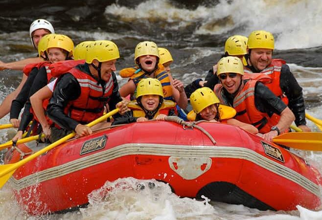 A group of people wearing helmets and life jackets paddle a red inflatable raft through rough whitewater rapids.