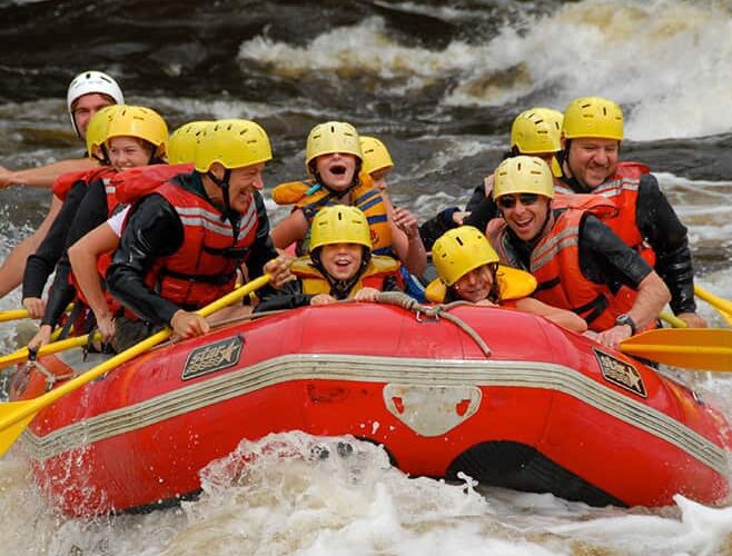 A group of people wearing helmets and life jackets paddle a red inflatable raft through rough whitewater rapids.