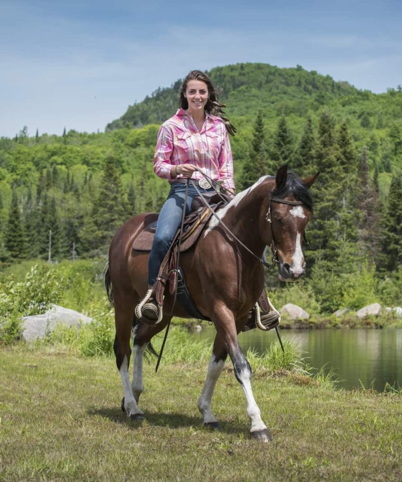 A person wearing a pink plaid shirt rides a brown and white horse on a grassy area near a pond, with trees and a hill in the background.