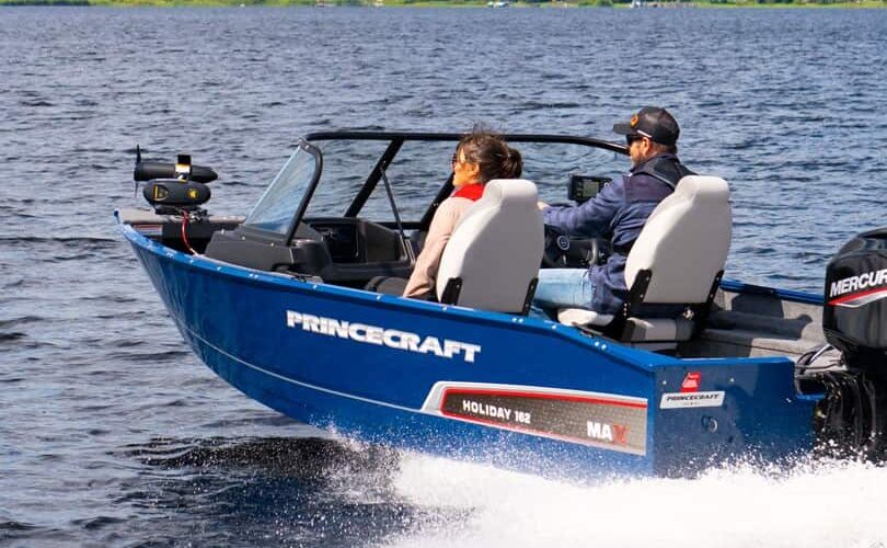 Two people ride a blue Princecraft Holiday 162 motorboat across a lake, with trees and houses visible on the far shore. Two people ride a blue Princecraft Holiday 162 motorboat across a lake, with trees and houses visible on the far shore.