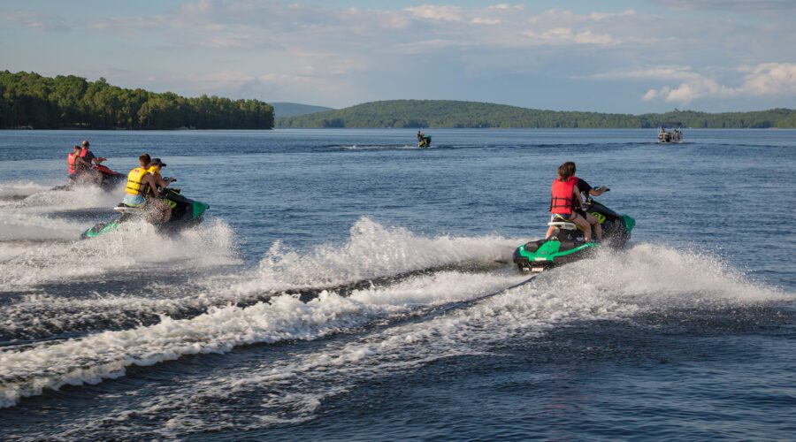 People riding jet skis on a large lake, wearing life jackets, with trees and hills in the background on a sunny day. People riding jet skis on a large lake, wearing life jackets, with trees and hills in the background on a sunny day.