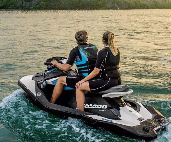 Two people wearing life jackets ride a jet ski on calm water near a treeline at sunset.