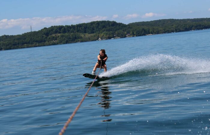 A person wakeboarding on a lake, holding a tow rope, with a forested shoreline and blue sky in the background.