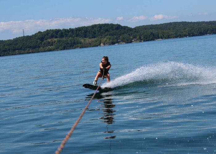 A person wakeboarding on a lake, holding a tow rope, with a forested shoreline and blue sky in the background.