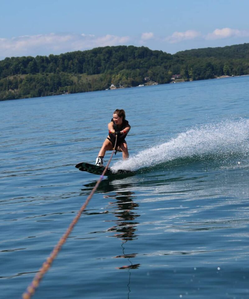 Une personne fait du wakeboard sur un lac calme, en pratiquant des sports nautiques tout en se tenant à une corde de remorquage, avec un rivage boisé et un ciel bleu en arrière-plan.