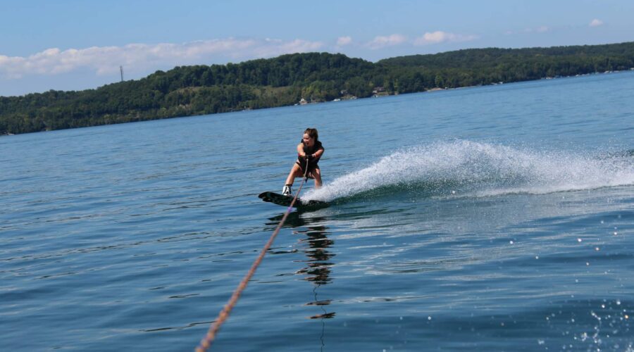 Une personne fait du wakeboard sur un lac calme, en pratiquant des sports nautiques tout en se tenant à une corde de remorquage, avec un rivage boisé et un ciel bleu en arrière-plan. Une personne fait du wakeboard sur un lac calme, en pratiquant des sports nautiques tout en se tenant à une corde de remorquage, avec un rivage boisé et un ciel bleu en arrière-plan.
