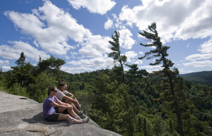 Two people sit on a rocky ledge overlooking a forested valley under a partly cloudy sky with tall trees nearby.