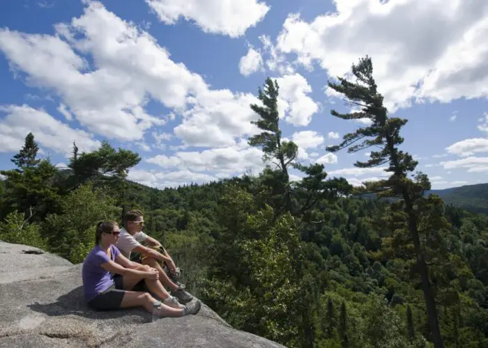 Two people sit on a rocky ledge overlooking a forested valley under a partly cloudy sky with tall trees nearby.