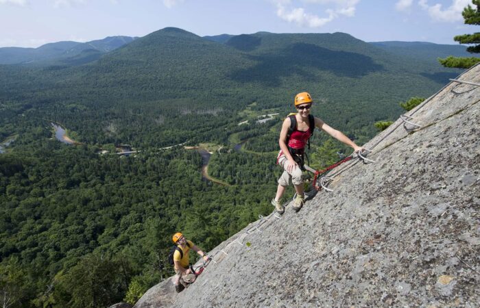 Two people wearing helmets and harnesses climb a steep rock face with aid of metal rungs; forested mountains and winding river are visible in the background.