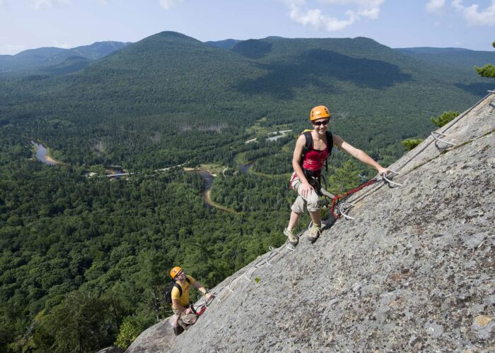 Two people wearing helmets and harnesses climb a steep rock face with aid of metal rungs; forested mountains and winding river are visible in the background.