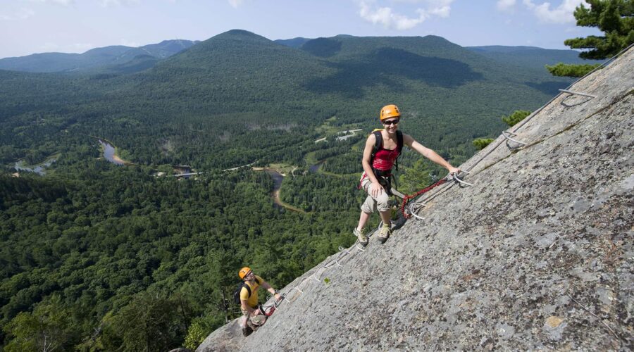 Deux personnes portant des casques et des harnais escaladent une paroi rocheuse abrupte sur la Via Ferrata Mont-Tremblant, à l’aide d’échelons métalliques, avec un paysage forestier et une route sinueuse en arrière-plan – une aventure dans un paysage époustouflant. Deux personnes portant des casques et des harnais escaladent une paroi rocheuse abrupte sur la Via Ferrata Mont-Tremblant, à l'aide d'échelons métalliques, avec un paysage forestier et une route sinueuse en arrière-plan - une aventure dans un paysage époustouflant.