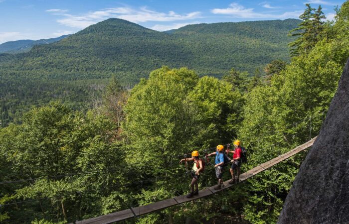 Three people wearing helmets walk across a narrow suspension bridge over a forested valley with mountains in the background.