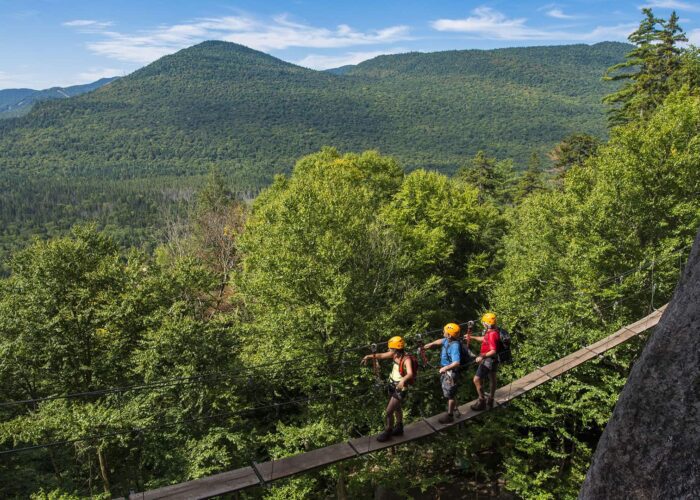 Trois personnes portant des casques et des harnais traversent un pont suspendu étroit dans une zone de montagne boisée sous un ciel bleu, profitant de l'une des nombreuses activités estivales offertes par la région.