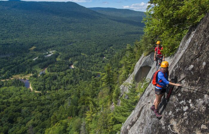Two people in helmets and harnesses climb a steep rock face, secured by cables, with a forested valley and winding river visible below.