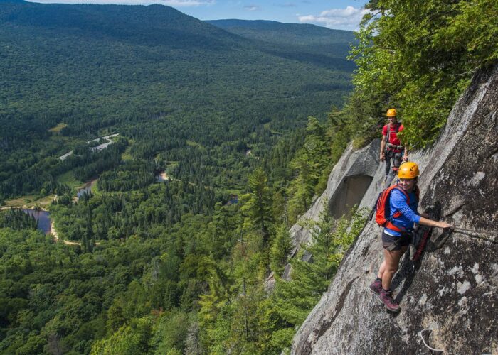 Two people in helmets and harnesses climb a steep rock face, secured by cables, with a forested valley and winding river visible below.