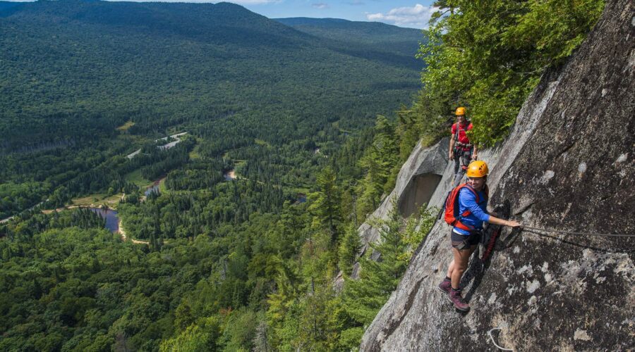 Deux personnes équipées de casques et de harnais escaladent une paroi rocheuse abrupte sur la Via Ferrata Mont-Tremblant, à l’aide de câbles de sécurité, en surplombant une vallée boisée et des montagnes. Deux personnes équipées de casques et de harnais escaladent une paroi rocheuse abrupte sur la Via Ferrata Mont-Tremblant, à l'aide de câbles de sécurité, en surplombant une vallée boisée et des montagnes.