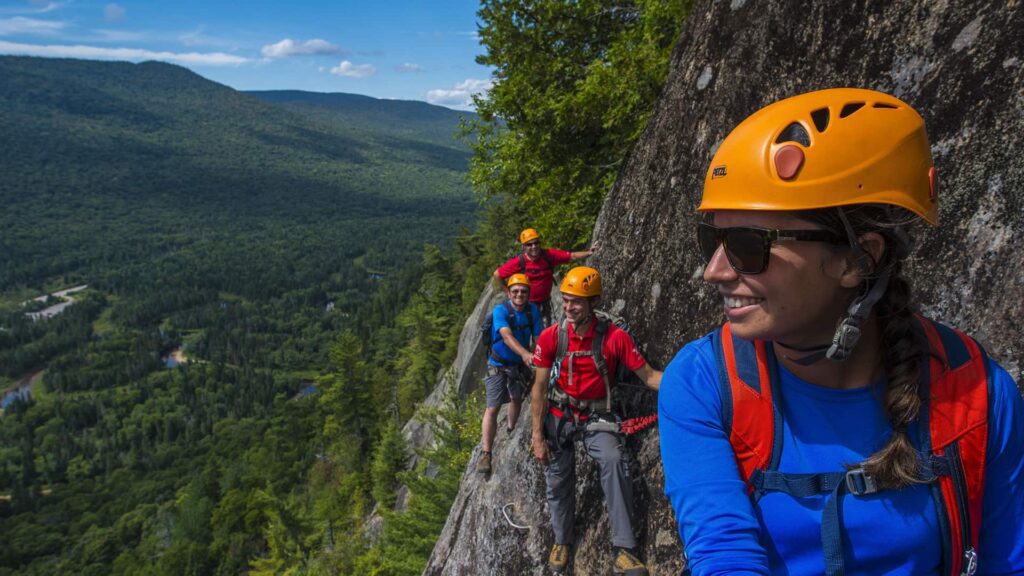 Four people wearing helmets and harnesses climb along a rocky mountain ledge with a forested valley and mountains in the background.