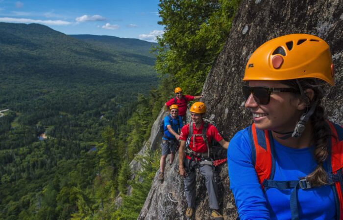 Four people wearing helmets and harnesses climb along a rocky mountain ledge with a forested valley and mountains in the background.