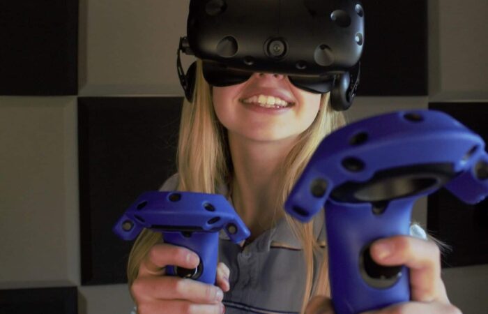 A person wearing a virtual reality headset and holding two blue VR controllers stands in front of a wall with black and gray square panels.