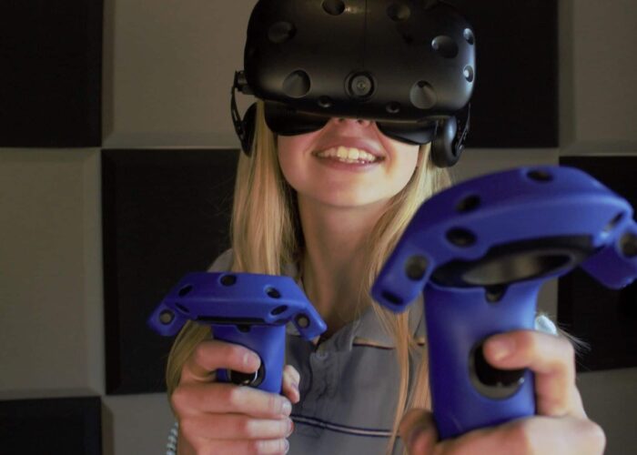 A person wearing a virtual reality headset and holding two blue VR controllers stands in front of a wall with black and gray square panels.