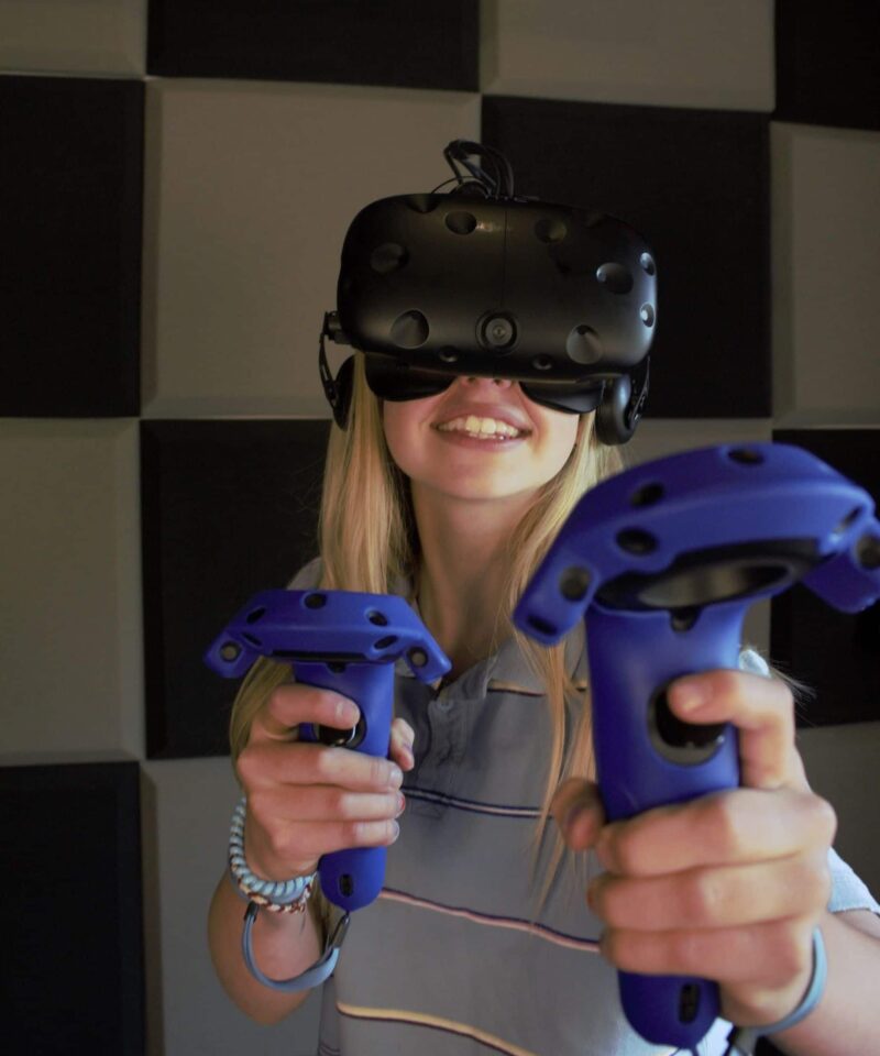 Une personne portant un casque de réalité virtuelle sourit tout en tenant deux contrôleurs VR bleus, debout devant un mur avec des panneaux acoustiques noirs dans une station de jeu Arcade RV.