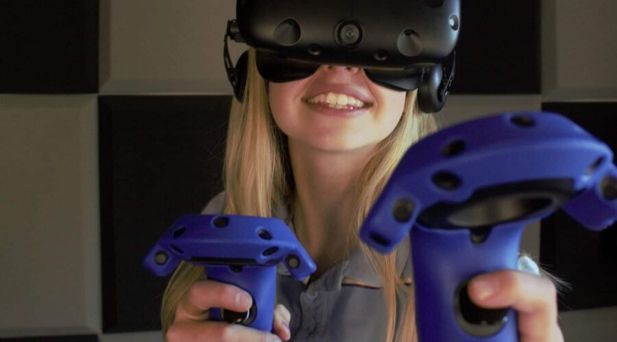 A person wearing a virtual reality headset and holding two blue VR controllers stands in front of a wall with black and gray square panels. A person wearing a virtual reality headset and holding two blue VR controllers stands in front of a wall with black and gray square panels.