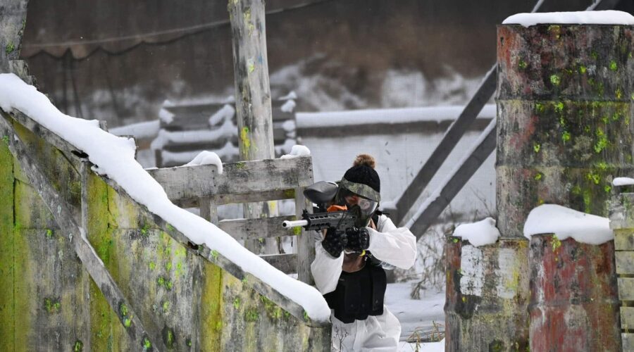 A person in winter gear and a protective mask aims a paintball gun while taking cover behind snow-covered barriers in an outdoor paintball field. A person in winter gear and a protective mask aims a paintball gun while taking cover behind snow-covered barriers in an outdoor paintball field.