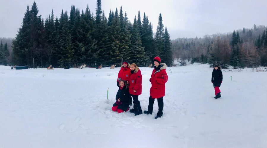 Four people dressed in red winter coats stand and kneel on snow, with a forest in the background; another person stands apart to the right.