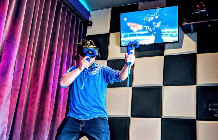 A person wearing a VR headset and holding controllers plays a virtual reality game in a room with padded walls and a monitor displaying gameplay.