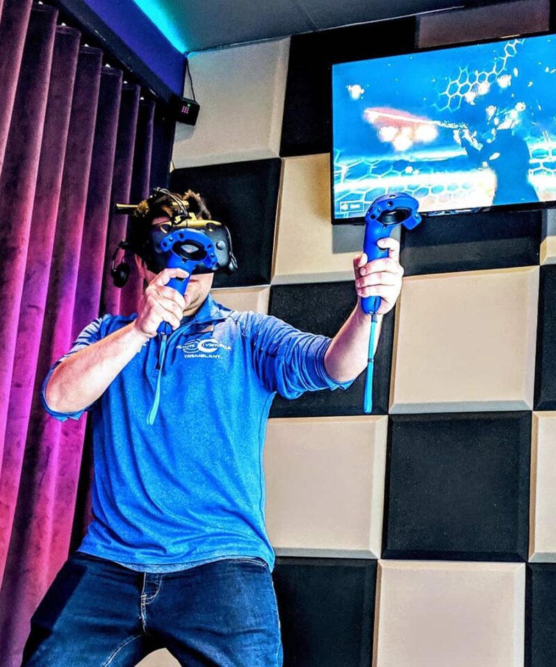 A person wearing a VR headset and holding controllers plays a virtual reality game in a room with padded walls and a monitor displaying gameplay.