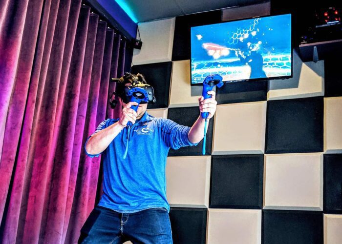 A person wearing a VR headset and holding controllers stands in a room with padded walls; a screen above displays virtual content.