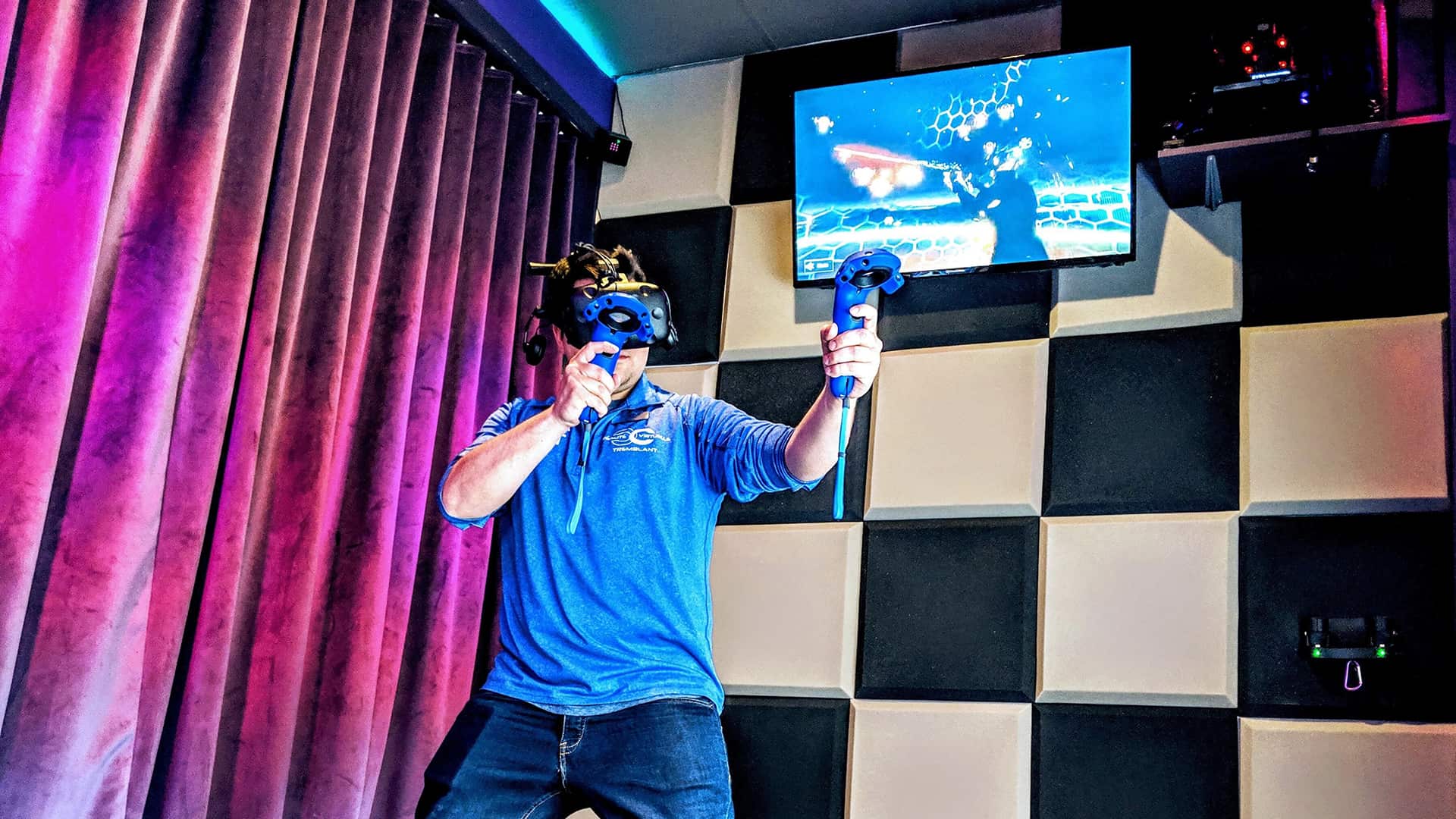 A person wearing a VR headset and holding controllers stands in a room with padded walls; a screen above displays virtual content.