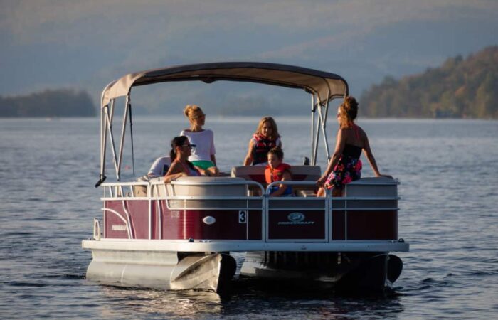A group of people, including children and adults, sit and stand on a pontoon boat on a calm lake with distant hills in the background.