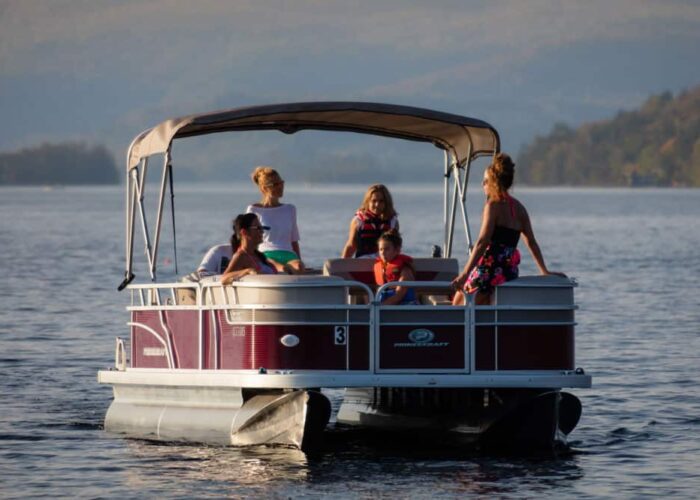 A group of people, including children and adults, sit and stand on a pontoon boat on a calm lake with distant hills in the background.