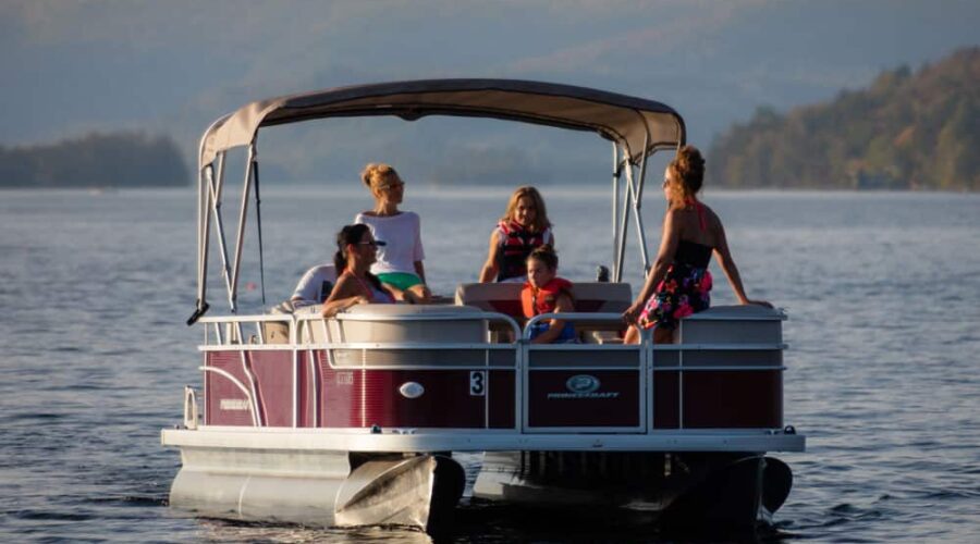 A group of people, including children and adults, sit and stand on a pontoon boat on a calm lake with distant hills in the background. A group of people, including children and adults, sit and stand on a pontoon boat on a calm lake with distant hills in the background.