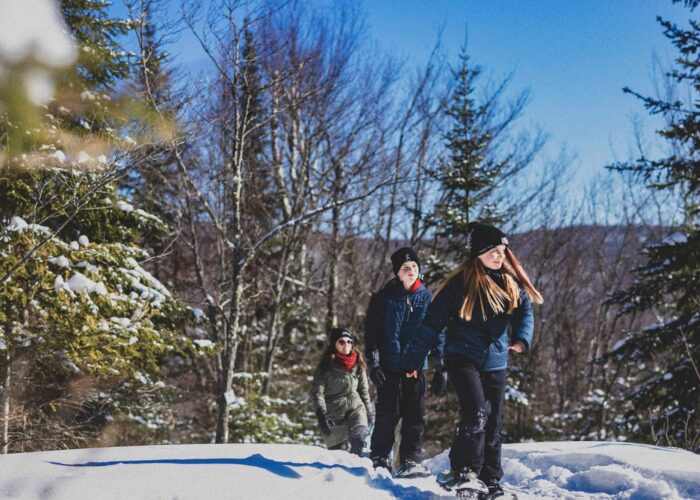 Three people dressed in winter clothing walk through a snowy forest trail on a sunny day, surrounded by snow-covered trees.