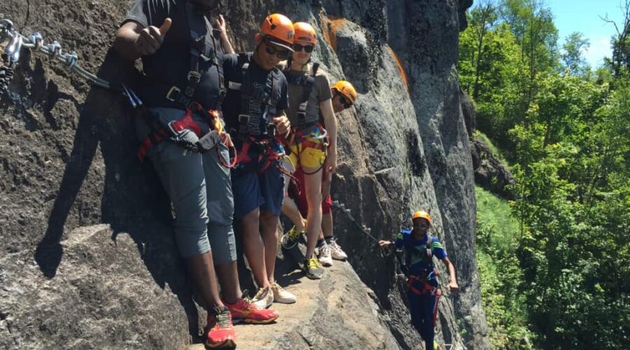 Five people wearing helmets and harnesses stand on a narrow rocky ledge attached to safety cables, participating in a rock climbing or via ferrata activity outdoors.