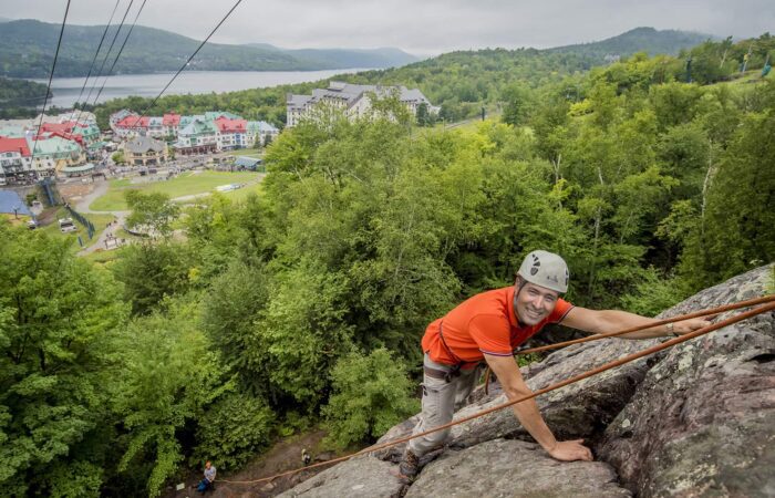 A man in a helmet and orange shirt rock climbing on a cliff face, with a resort town, lake, and forested hills in the background.
