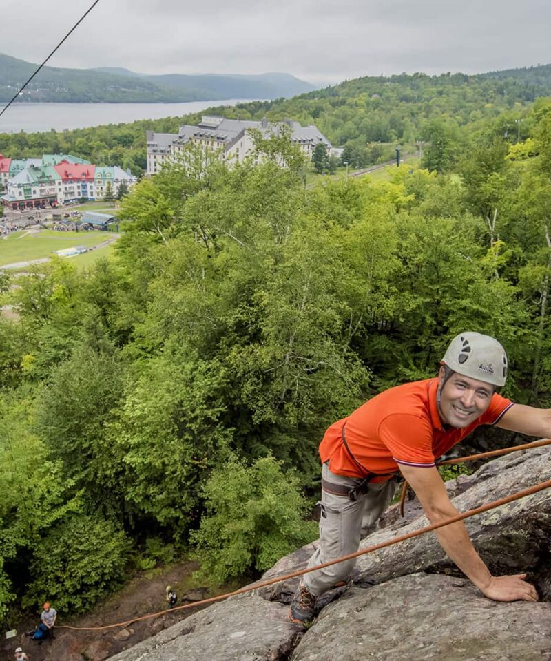 A man in a helmet and orange shirt rock climbing on a cliff face, with a resort town, lake, and forested hills in the background.