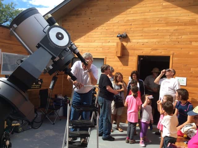 A group of people, including children, gather outside a wooden building as a man uses a large telescope while others watch or use small devices to observe.