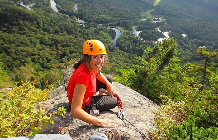 A woman in a helmet and climbing gear sits on a rocky ledge, smiling, with a forested valley and winding river visible in the background.