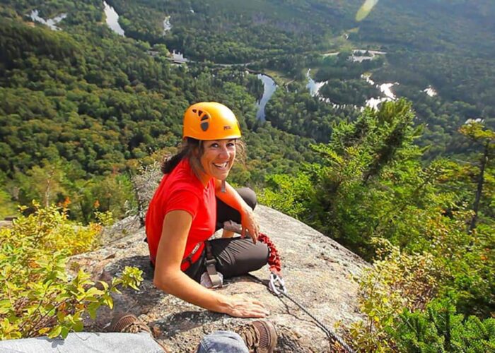 A woman in a helmet and climbing gear sits on a rocky ledge, smiling, with a forested valley and winding river visible in the background.