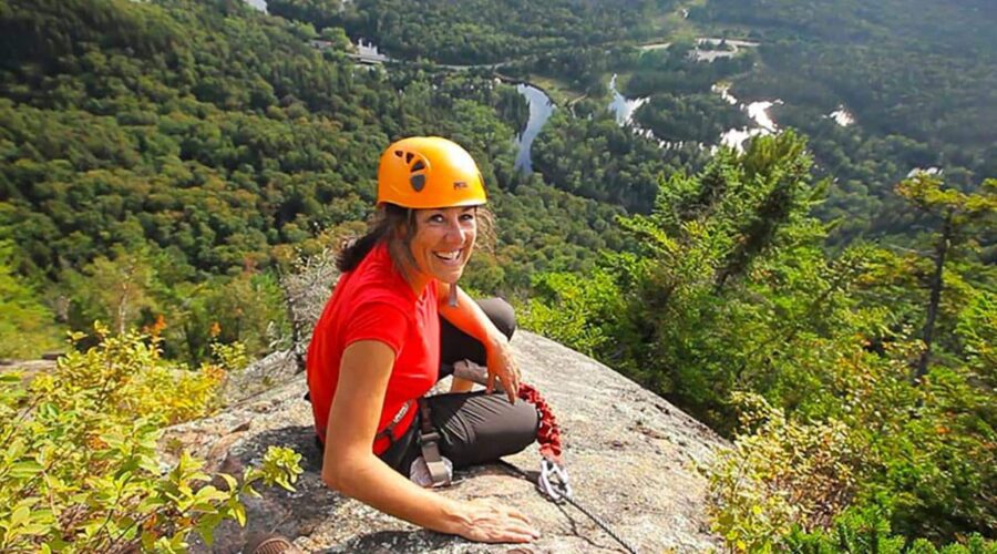 Une femme portant un casque et un harnais est assise sur une corniche rocheuse le long de la Via Ferrata Mont-Tremblant, surplombant un paysage forestier avec des rivières en contrebas à coucher du soleil. Une femme portant un casque et un harnais est assise sur une corniche rocheuse le long de la Via Ferrata Mont-Tremblant, surplombant un paysage forestier avec des rivières en contrebas à coucher du soleil.