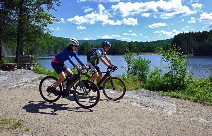 Deux cyclistes casqués font du vélo électrique sur un chemin de gravier au bord d'un lac au Domaine Saint-Bernard, entouré d'arbres et de collines sous un ciel bleu avec des nuages épars - parfait pour les adeptes de la location de vélos.