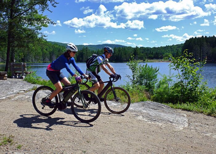 Two cyclists ride on a dirt path beside a lake, surrounded by trees and greenery under a blue sky with scattered clouds.