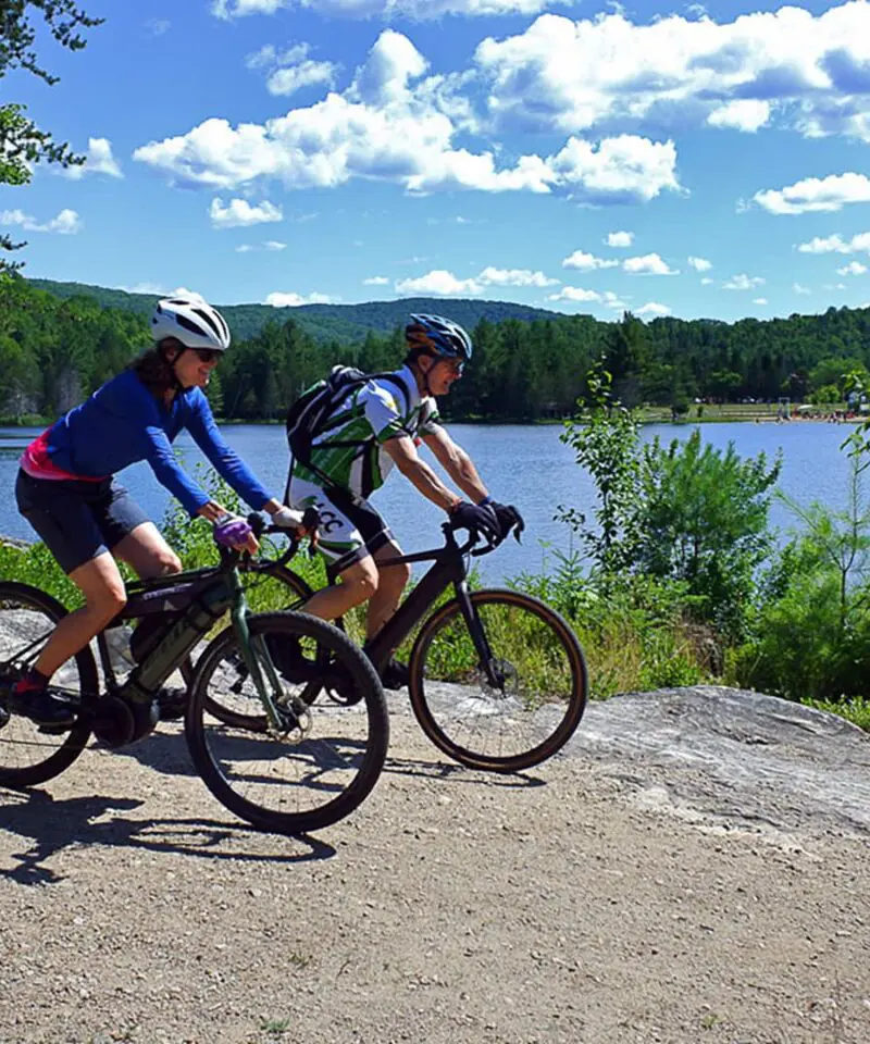 Two cyclists ride on a dirt path beside a lake, surrounded by trees and greenery under a blue sky with scattered clouds.