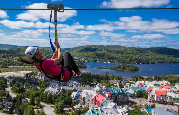 A person wearing a helmet rides a zipline above a town with colorful rooftops, surrounded by green hills and a lake under a partly cloudy sky. A person wearing a helmet rides a zipline above a town with colorful rooftops, surrounded by green hills and a lake under a partly cloudy sky.