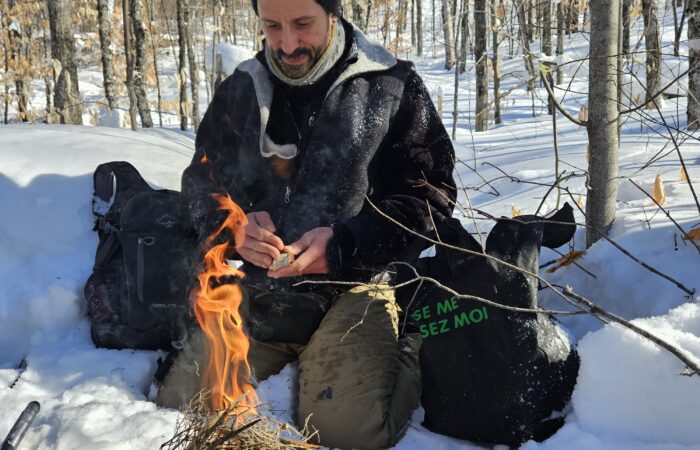 A man, known as the Fire Man, sits in the snow in a forest, tending to a campfire made of sticks. Clad in winter gear and surrounded by bags and leafless trees, he prepares for an adventurous snowshoeing guided tour under a clear blue sky.