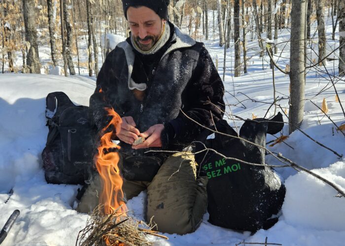 A man, known as the Fire Man, sits in the snow in a forest, tending to a campfire made of sticks. Clad in winter gear and surrounded by bags and leafless trees, he prepares for an adventurous snowshoeing guided tour under a clear blue sky.